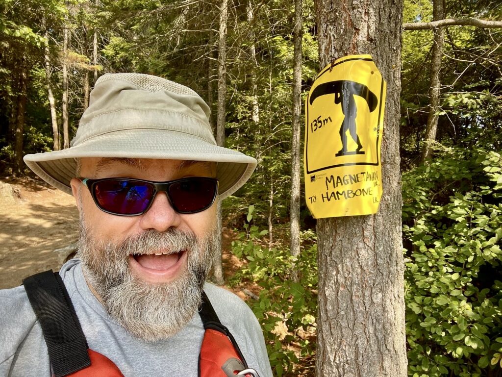 Middle aged man wearing sunglasses a tilley hat standing in front of a tree with a yellow portage sign reading Magnetewan to Hambone